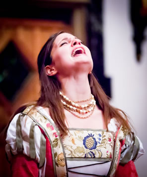 Juliet in red dress with white bodice embroidered with colorful flowers, cut slives of silver and white, and three chokers of pearls has her head back as she shouts.