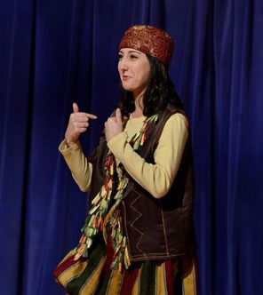 Grumio in red leather servants cap, gold, red, and green striped renaissance pats, bown leather vest with multicolor tassles points at herself with both hands.