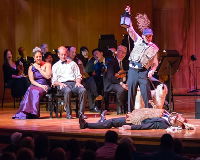 Hippolyta in purple gown and crown sits next to Theseus in white suit shirt and bowtie in front of members of the orchestra string section, all in black formal wear, leaning over to see, bottom in whicker breastplate and funny helmet lies on the floor as Moonshine in flack-jacket, baseball cap with thorn bush, holding up a lantern and leash on white dog stands over him.