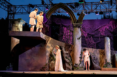 Production photo of Arcite and Paloman up on the ledge looking down at Emilia in the garden below, her attendant in the backgorund.