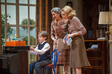 Young Ethan in private school uniform works a video player on top of a TV as Jean and Hester watch, Jean holding a tablet, Hester holding the video box, witht he patio in the background