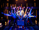 Vy, wearing a jellow jacket, white blouse, orange patterend dress, white socks, plain shoes, and a wide brim hat stands with both hands holding a battered suticase in front of her as the ensemble behind her leans out with outstreched hands creating a starburst effect. Photo by Daniel Rader