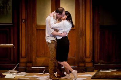 Winston and Julia kiss, he in white shirt and brown pants, she in white shirt and knee-length black skirt and white bobby sox, with clutter all about the office canteen