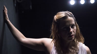 Gilbert with right hand on the wall and disheveled hair looks off to her left against a black background with three spotlights above.