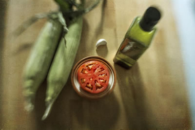 Tomato cut in half and in bowl in center of wood cutting board; arranged around it left to right, two corn cobs still sheathed, garlic clove, bottle of olive oil